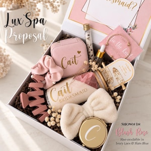 May include: A white gift box containing a pink jewelry case labeled Cait, a pink scrunchie, a pink hair claw, a glass vial of bath salts, a gold bangle, a hair clip, a tumbler labeled Cait Bridesmaid, a white headband, and a gold compact mirror.
