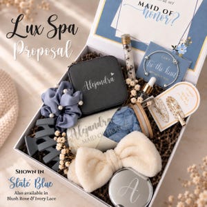 May include: A gift box containing a black jewelry case labeled Alejandra, a slate blue scrunchie with pearls, a slate blue hair claw, a glass jar with blue petals labeled Alejandra Maid of Honor, a cream headband, a mirror, and hair accessories.