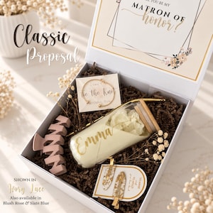 May include: A white gift box containing a glass tumbler with a bamboo lid and the name Emma, a gold-toned bracelet on a card reading I can't tie the knot without you, two hair clips with gold flakes, and a matte mauve hair claw on brown paper filler.