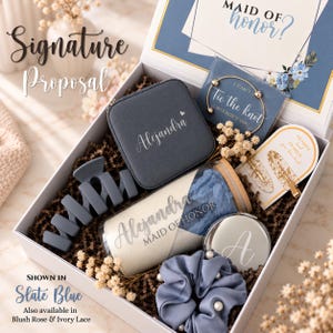 May include: A white gift box containing a slate blue jewelry case personalized with the name Alejandra, a glass tumbler with a bamboo lid, a slate blue hair claw, a pearl-accented scrunchie, a gold-tone bracelet, a compact mirror, and hair clips.