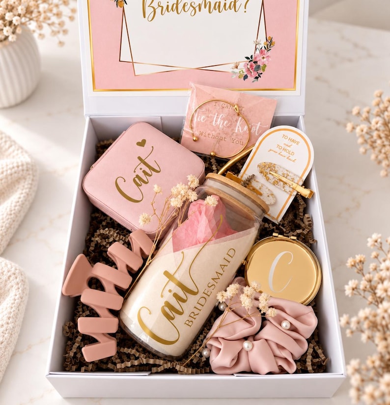 May include: A white gift box containing a pink jewelry case labeled Cait, a glass tumbler with a bamboo lid labeled Cait Bridesmaid, a gold bracelet, a hair clip on a card, a round gold compact mirror with a C, and a pink pearl-embellished scrunchie.