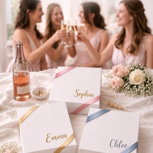 May include: Three white square gift boxes with satin ribbons in pink, gold, and blue. Each box features a name printed in gold or silver script: Sophia, Emma, and Chloe. The boxes are arranged on a white fabric surface with small floral accents.