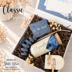 May include: A gift box containing a glass tumbler with a bamboo lid and straw, a slate blue hair claw clip, a silver-toned bangle bracelet on a card reading 'I can't Tie the Knot Without You', and two gold-flecked hair clips on a display card.