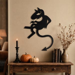 May include: A black metal wall art piece shaped like a stylized cartoon devil with horns and a pointed tail. The figure is depicted in a dynamic pose with one arm raised and legs bent, featuring a smooth matte finish and clean, sharp edges.