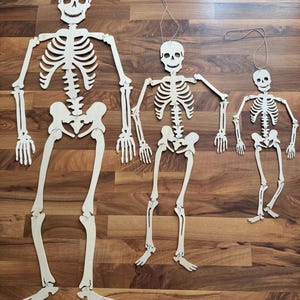 May include: Three wooden skeleton decorations of varying sizes, each with a string for hanging. The skeletons are light beige and have detailed rib cages, skulls, and articulated limbs. They are arranged on a brown wooden surface.