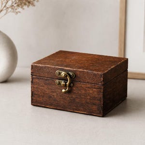 May include: A rectangular wooden box with a dark brown stained finish and visible wood grain. The box features a hinged lid secured by a brass-colored metal latch centered on the front panel. The construction shows small finger joints at the corners.