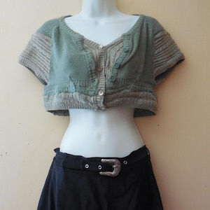 May include: A sage green and beige short-sleeved cropped cardigan with a button-down front, paired with a black mini skirt featuring a silver-toned western-style belt buckle and a layered, frayed pink and black hemline detail.