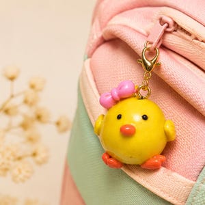 May include: A yellow chick charm with a pink bow, orange beak, and orange feet, attached to a gold-toned lobster clasp. The charm is hanging from the zip pull of a pastel pink and light green fabric bag with a visible zip track.
