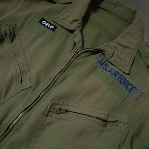 以下が含まれることがあります： オリーブグリーンのミリタリースタイルのジャンプスーツ、ジッパー開閉。襟、複数のポケット、そして「GRUP」と「U.S. AIR FORCE」の青い文字が入ったパッチが特徴です。ジャンプスーツはヴィンテージ感があります。