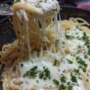 May include: Close-up of spaghetti with melted cheese and chopped parsley. The cheese stretches as a fork lifts a portion of the pasta. The dish is in a dark pan, with more pasta visible in the background. A delicious, creamy meal.