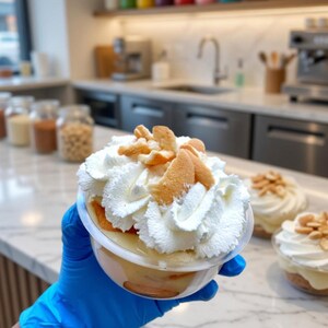 Könnte beinhalten: Ein klarer Plastikbecher gefüllt mit Bananenpudding, garniert mit Schlagsahne und Vanillewaffeln. Das Dessert wird von einer Person mit einem blauen Handschuh gehalten. Weitere Desserts sind im Hintergrund zu sehen.
