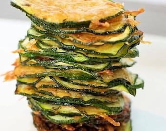 Può includere: Una pila di croccanti chips di zucchine, che mostra un mix di tonalità verdi e dorate. Le chips sono disposte a torre, con trame visibili e leggere bruciature sui bordi, suggerendo una preparazione fritta o al forno. L'immagine mette in risalto gli ingredienti freschi e naturali.