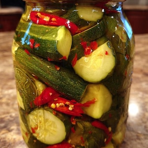 Könnte beinhalten: Ein klares Glas mit Gurkenscheiben und roten Chilischoten in Salzlake. Das Glas hat einen silbernen Deckel. Die Gurken sind grün und die Paprika leuchtend rot.