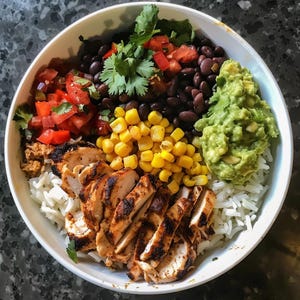 May include: A white bowl filled with a colorful assortment of food. Grilled chicken, white rice, black beans, corn, guacamole, and diced tomatoes are arranged in sections. Fresh cilantro adds a vibrant green touch to the dish.