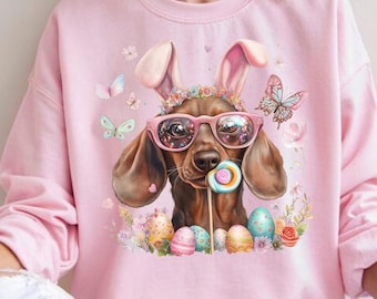 May include: A pink sweatshirt with a graphic of a dachshund wearing bunny ears, sunglasses, and a lollipop. The dachshund is surrounded by flowers and Easter eggs.