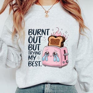 May include: A white sweatshirt with a pink toaster and a burnt piece of bread on top. The toaster has a sad face and is crying. The text on the sweatshirt reads "Burnt out but trying my best."