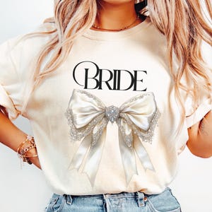 May include: A white t-shirt with the word "BRIDE" printed in black and a large white satin bow with a jeweled centre.