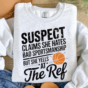 Könnte beinhalten: Ein graues Sweatshirt mit dem Text "SUSPECT CLAIMS SHE HATES BAD SPORTSMANSHIP BUT SHE YELLS AT THE REF" in Schwarz. Neben dem Text befindet sich eine Basketballgrafik.