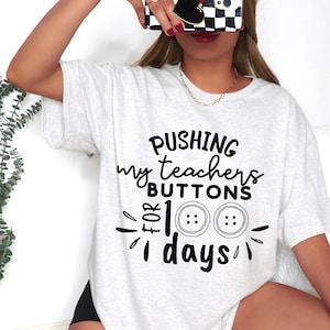 May include: A white t-shirt with black text that reads "Pushing my teachers buttons for 100 days". The text is surrounded by a black border and includes two black buttons.