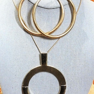 May include: Gold-toned jewelry set featuring large hoop earrings and a pendant necklace with a circular design. The necklace has a rectangular top piece. The jewelry is displayed on a light blue fabric.