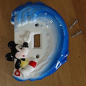 May include: A ceramic light switch cover featuring Mickey Mouse in a wave design. The cover is white with a blue wave and Mickey Mouse wearing a yellow shirt and red shorts. Includes two screws.