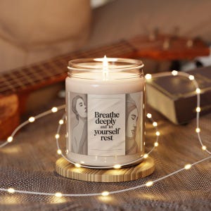 Pode incluir: Uma vela acesa num frasco de vidro com o texto "Breathe deeply and let yourself rest" num r&oacute;tulo com duas figuras femininas esbo&ccedil;adas. A vela est&aacute; sobre um descanso de copo redondo de madeira, com luzes de corda e uma guitarra no fundo.