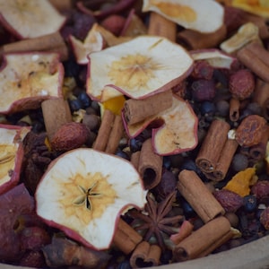 Op de afbeelding: Een close-up van een kom gevuld met gedroogd fruit, specerijen en sinaasappelschillen. Het mengsel bevat appelschijfjes, kaneelstokjes, steranijs en kruidnagels.
