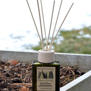 May include: A green glass bottle with a wooden lid and brown reeds. The bottle is labeled "Maine Woods Reed Diffuser" with the website address "www.mainescents.net" and the size "4 fl. oz/118.2 ml."