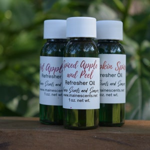 Puede incluir: Tres pequeñas botellas de vidrio verde con etiquetas blancas. Las etiquetas dicen "Hot Apple Refresher", "Spiced Apple and Peel Refresher Oil" y "Pumpkin Spice Refresher Oil". Todas las botellas están etiquetadas con "Maine Scents and Seasons" y una dirección de sitio web.