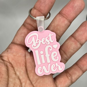 Puede incluir: Llavero rosa con una inscripción blanca que dice "Best life ever" con un pequeño símbolo de corazón.