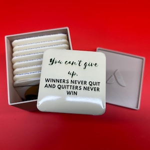 Puede incluir: Un posavasos cuadrado blanco con el texto "You can't give up. Winners never quit and quitters never win" en verde y negro. El posavasos está junto a una caja de posavasos similares, todo sobre un fondo rojo.