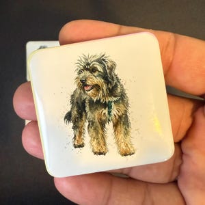 Puede incluir: Botón cuadrado y brillante con una ilustración en acuarela de un perro mestizo tipo terrier. El perro tiene pelaje negro y marrón, un collar verde y saca la lengua. El botón tiene un fondo crema y un borde rosa.