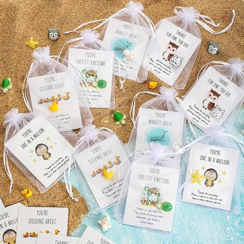 Pode incluir: Uma cole&ccedil;&atilde;o de pequenos sacos de organza brancos amarrados com fitas brancas, cada um contendo um pequeno presente e um cart&atilde;o. Os cart&otilde;es apresentam ilustra&ccedil;&otilde;es de animais e frases como "You're One in a Million" em uma superf&iacute;cie arenosa.
