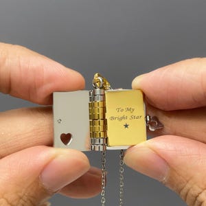 Könnte beinhalten: Eine silber- und goldfarbene Medaillon-Halskette in Form eines kleinen Buches. Das Medaillon ist geöffnet und zeigt eine goldfarbene Seite mit der Aufschrift "To My Bright Star" und einem Sternsymbol. Die linke Seite hat einen herzförmigen Ausschnitt.