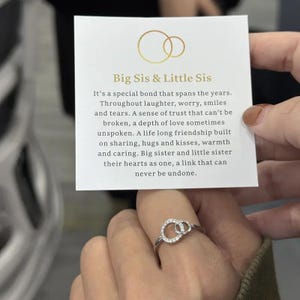 May include: A silver ring with two interlocking circles, each encrusted with small, clear stones, is displayed. The ring is on a finger, with a card in the background that reads "Big Sis & Little Sis" and describes a special bond.