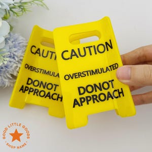May include: Two bright yellow plastic caution signs featuring a cutout handle at the top. The text printed in bold black letters reads CAUTION OVERSTIMULATED DONOT APPROACH. The signs are rectangular with a slight curve and notched bottom edges.