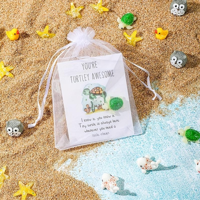 Pode incluir: Uma sacola de presente de organza branca com um cart&atilde;o que diz "You're Turtley Awesome" com uma ilustra&ccedil;&atilde;o de tartaruga. A sacola &eacute; cercada por areia, estrelas do mar e pequenas figuras de tartarugas e patos.