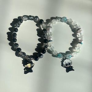 May include: Two beaded bracelets. One bracelet features black and clear beads with a black charm. The other bracelet has white and clear beads with a blue charm. Both bracelets are on a white surface.