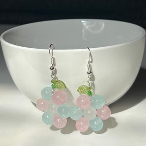 May include: A pair of handmade earrings featuring clusters of pink and light blue beads, resembling grapes. Each earring has a silver hook and a small green leaf accent. The earrings are displayed in front of a white bowl.