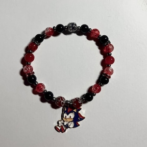 May include: A beaded bracelet featuring red and black beads with a silver skull accent. A blue, red, and white cartoon character charm hangs from the bracelet. The bracelet is on a white surface.