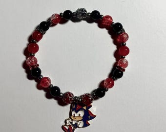 May include: A beaded bracelet featuring red and black beads with a silver skull accent. A blue, red, and white cartoon character charm hangs from the bracelet. The bracelet is on a white surface.