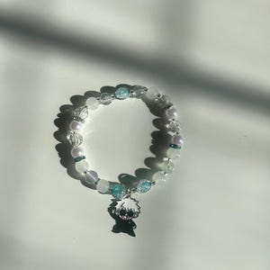May include: A beaded bracelet featuring white, clear, and light blue beads. The bracelet includes a charm with a black and white anime character. The bracelet is on a white surface.