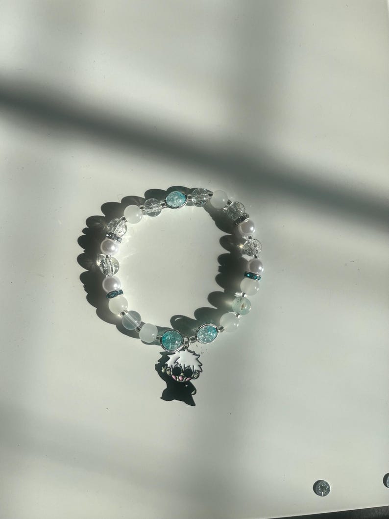 May include: A beaded bracelet featuring white, clear, and light blue beads. The bracelet includes a charm with a black and white anime character. The bracelet is on a white surface.