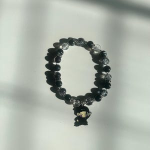 May include: Black beaded bracelet with a charm. The bracelet features alternating black and clear beads. A small charm of a cartoon character is attached to the bracelet.