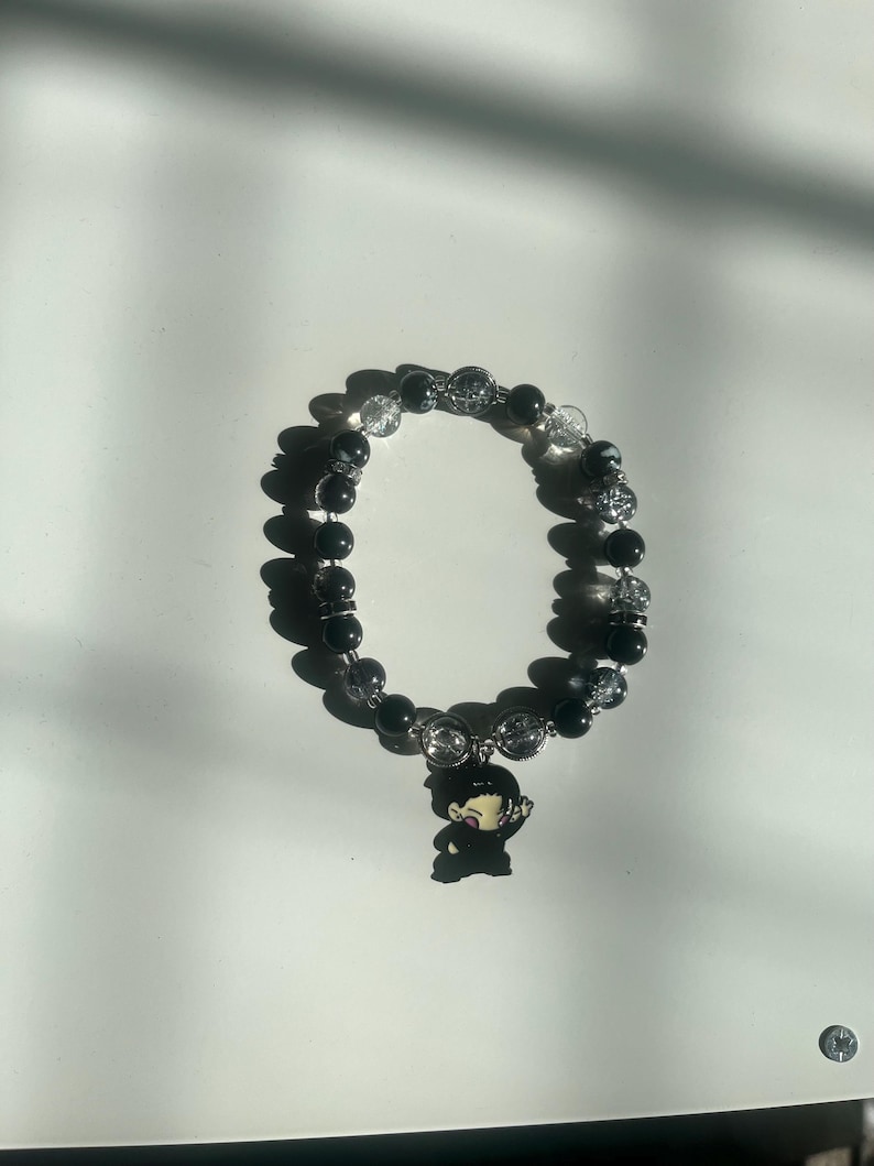 May include: Black beaded bracelet with a charm. The bracelet features alternating black and clear beads. A small charm of a cartoon character is attached to the bracelet.