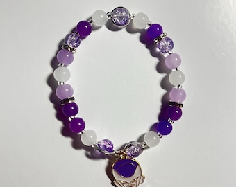 May include: A beaded bracelet featuring a mix of purple, lavender, and white beads. The bracelet has a charm with a purple design and silver accents. The beads are strung together with a silver-colored elastic cord.