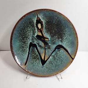 May include: A round ceramic plate featuring a mottled teal and black glaze with a speckled texture. The center displays a dark, abstract botanical motif resembling a cattail or plant stalk, with a brown rim encircling the edge.