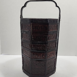 May include: A dark brown, hexagonal, tiered food carrier with four stacked compartments and a tall, arched handle. The structure features woven panels framed by smooth, solid borders, creating a vertical, multi-level storage container.
