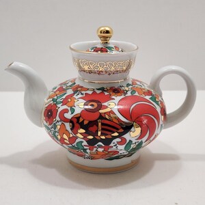 May include: A white porcelain teapot featuring a vibrant red, orange, and black rooster motif surrounded by floral patterns. The teapot includes a matching lid with a gold-tone knob, gold-trimmed edges, and a curved handle and spout.