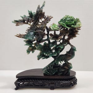 May include: A carved green and dark brown stone sculpture depicting two birds perched among leafy branches and large flower blossoms. The intricate stone carving is mounted on a dark, polished wooden base with ornate, carved feet.